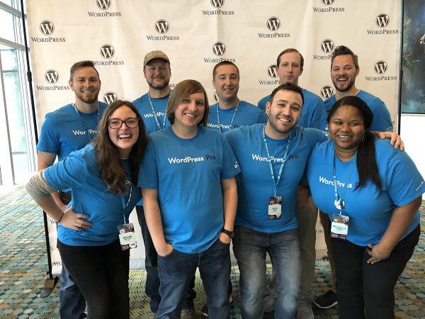 9 Bluehost employees at WordCamp US 2018 posing in front of a photo backdrop with the WordPress logo. All are wearing Bluehost blue colored shirts.