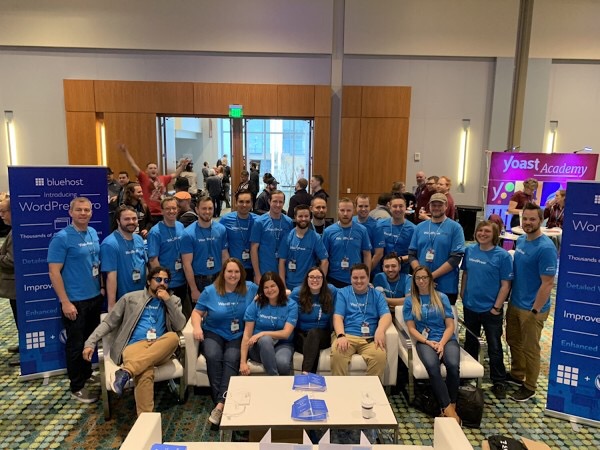 The Bluehost team posing for a group photo in the company's booth at WordCamp US 2018. Some are sitting on a couch in the front and the rest are standing in the back. All are wearing Bluehost blue colored shirts.