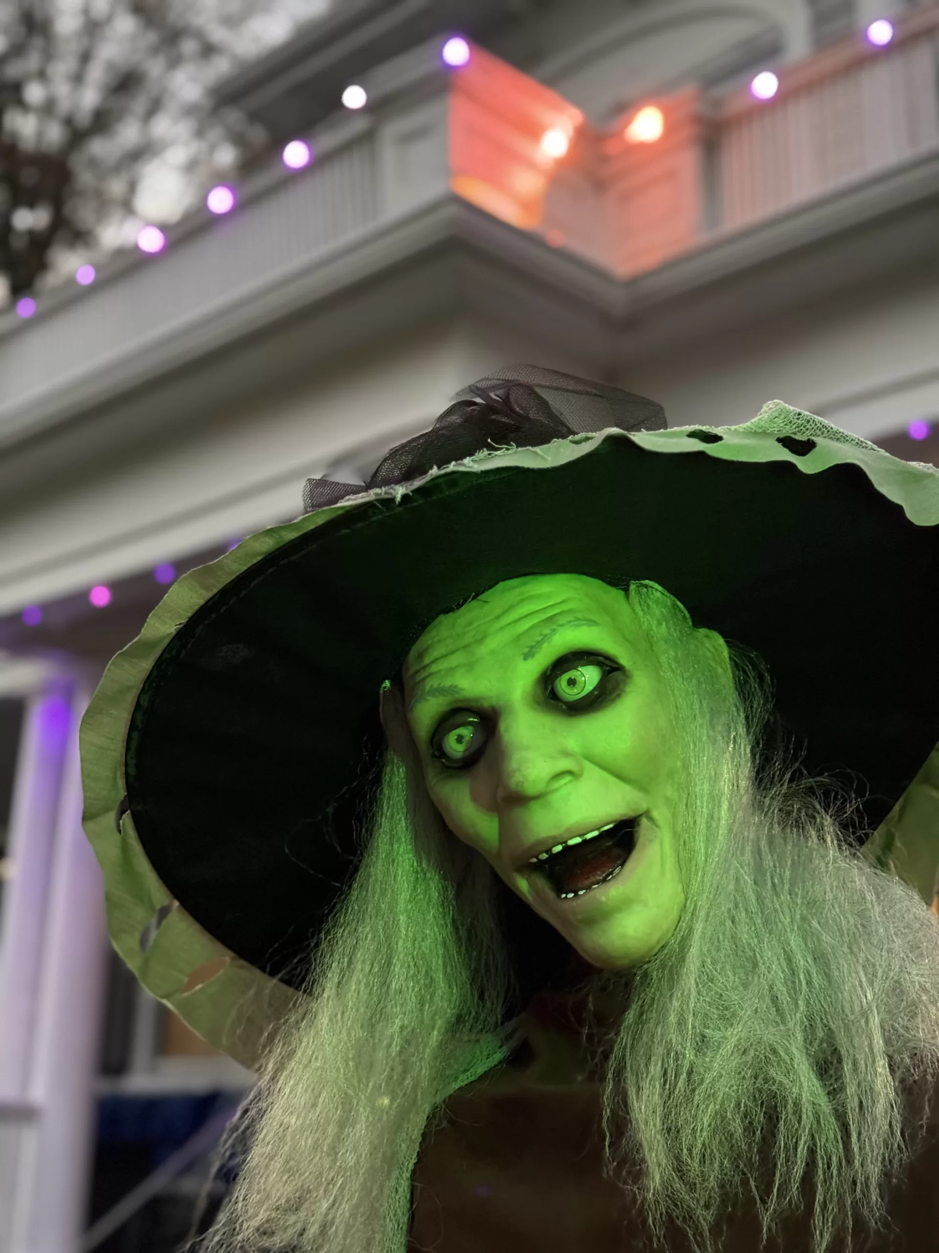 A witch mannequin in the foreground as part of Halloween decorations. The house is out of focus in the background, but decorative lights can be seen outlining the house.