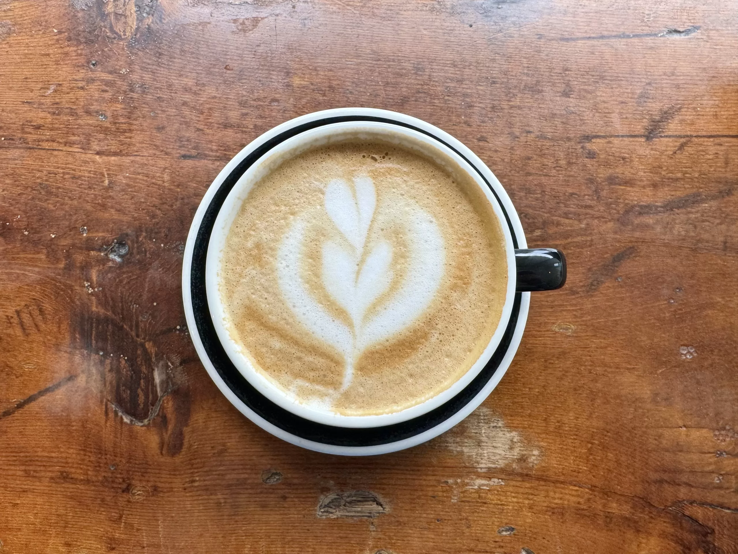 An overhead view of a cappuccino in a mug with milk art on a worn, dark wooden counter.