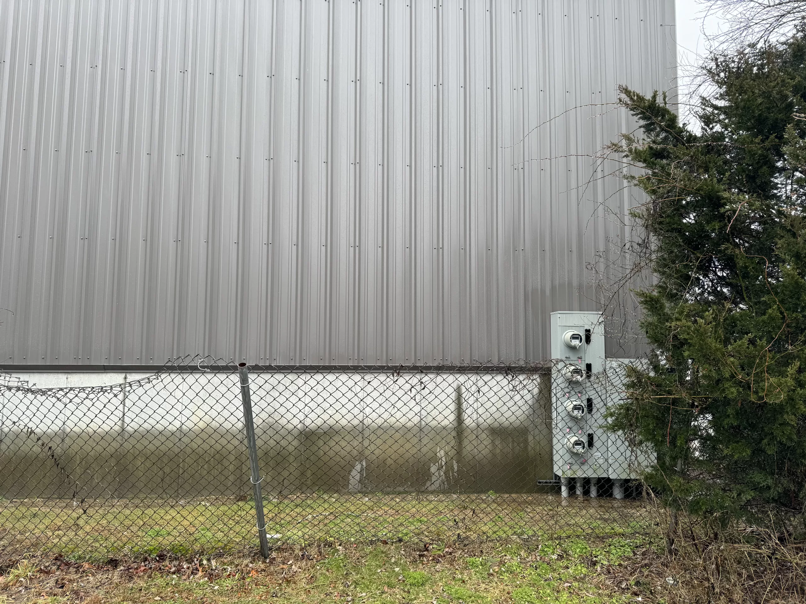 The side of a grey industrial building on a rainy day with a falling down chain link fence in front. 