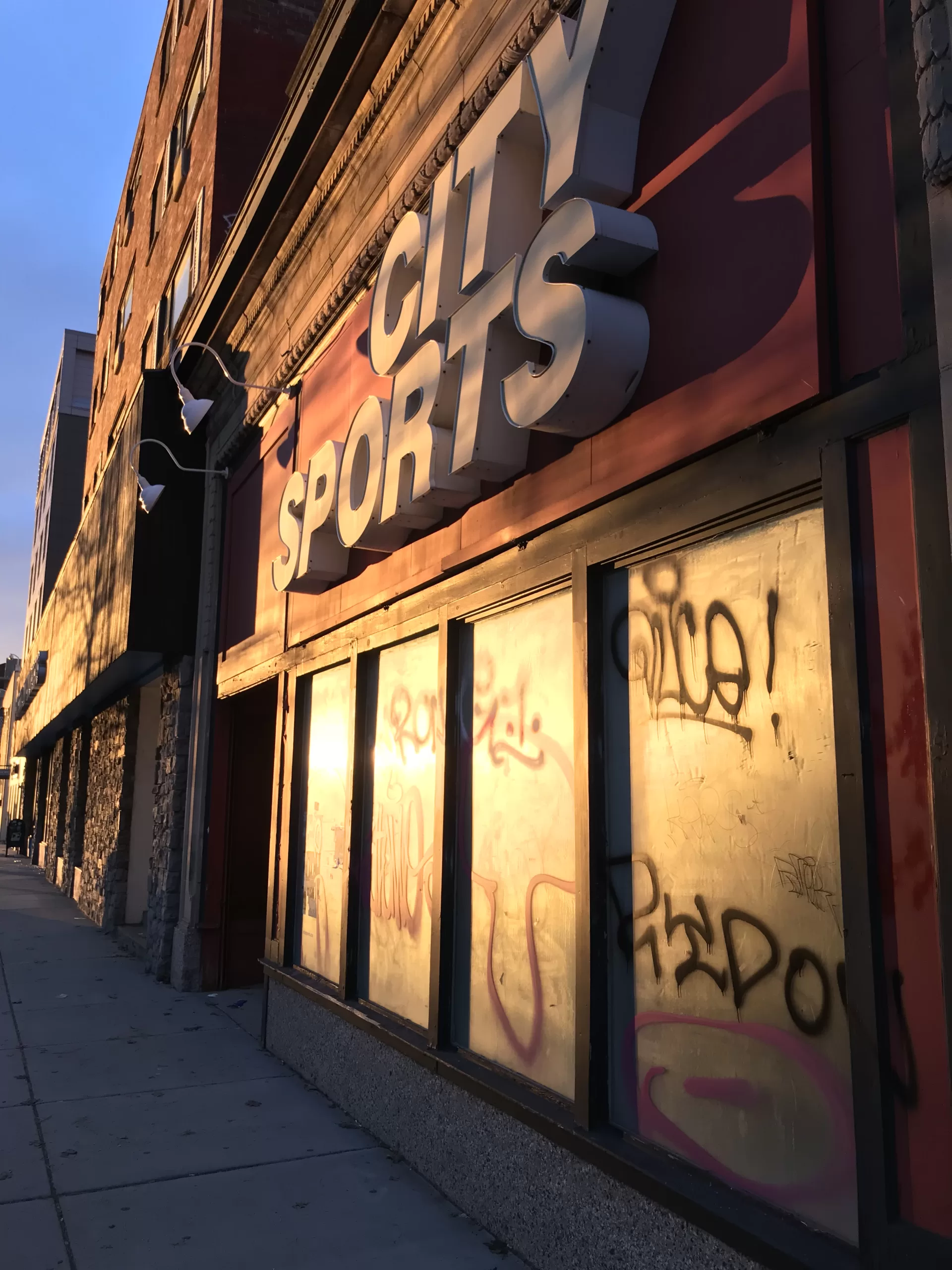 An abandoned City Sports store front on Commonwealth Avenue in North Brookline, MA. The windows are covered on the inside and there's graffiti on the outside. The sun is setting and shining warm light on the building.