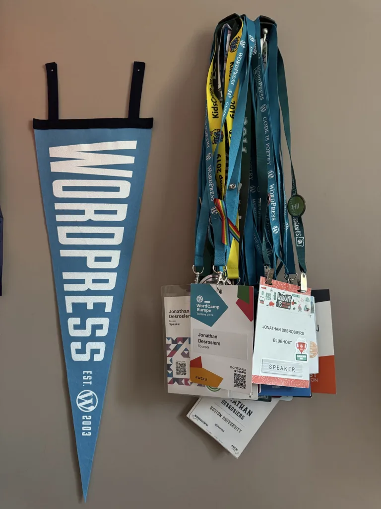 Dozens of WordCamp and conference badges hanging on a wall hook. A WordPress pennant is hung on the wall next to them.