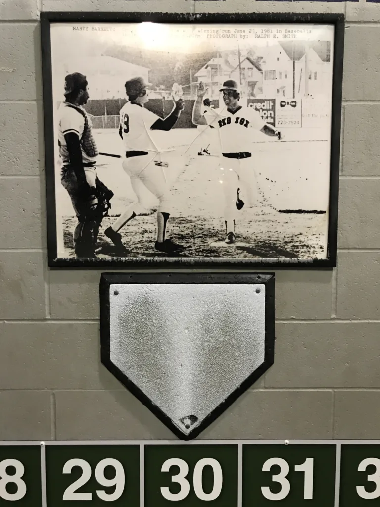 A home plate bolted to a white cinder block wall above the full box score from the longest game in the history of professional baseball. Above the plate is a photo of Marty Barrett touching the plate to score the winning run.