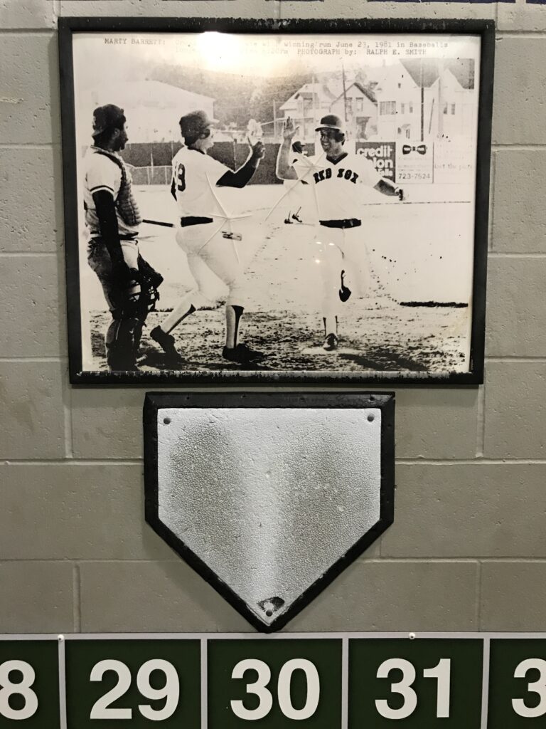 A home plate bolted to a white cinder block wall above the full box score from the longest game in the history of professional baseball. Above the plate is a photo of Marty Barrett touching the plate to score the winning run.