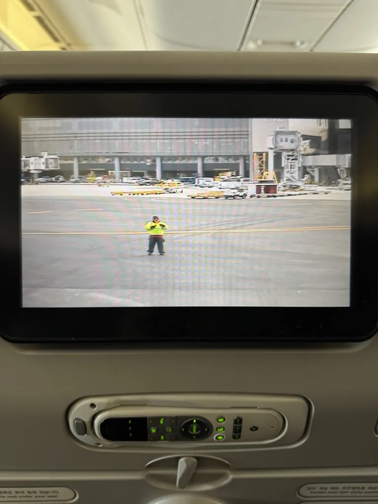 The seat back screen of an airplane seat showing the front facing camera on the aircraft. An airport worker can be seen holding batons in a cross position.