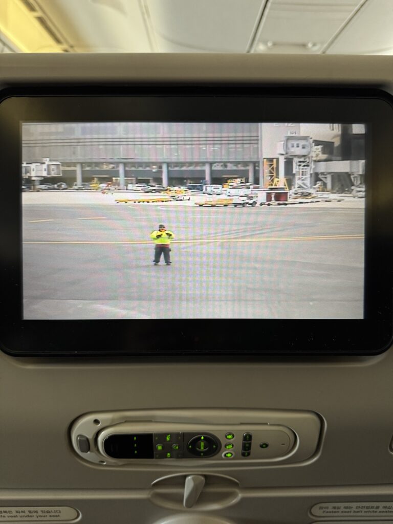 The seat back screen of an airplane seat showing the front facing camera on the aircraft. An airport worker can be seen holding batons in a cross position.