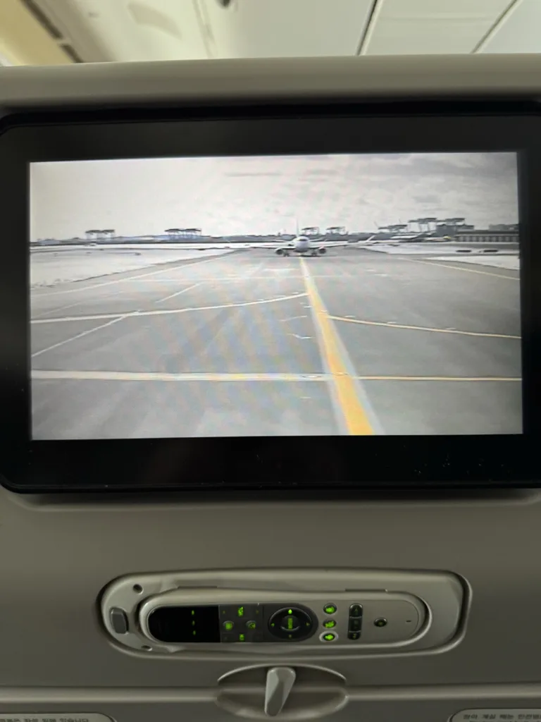 The seat back screen of an airplane seat showing the front facing camera on the aircraft. Another airplane can be seen queued in front.