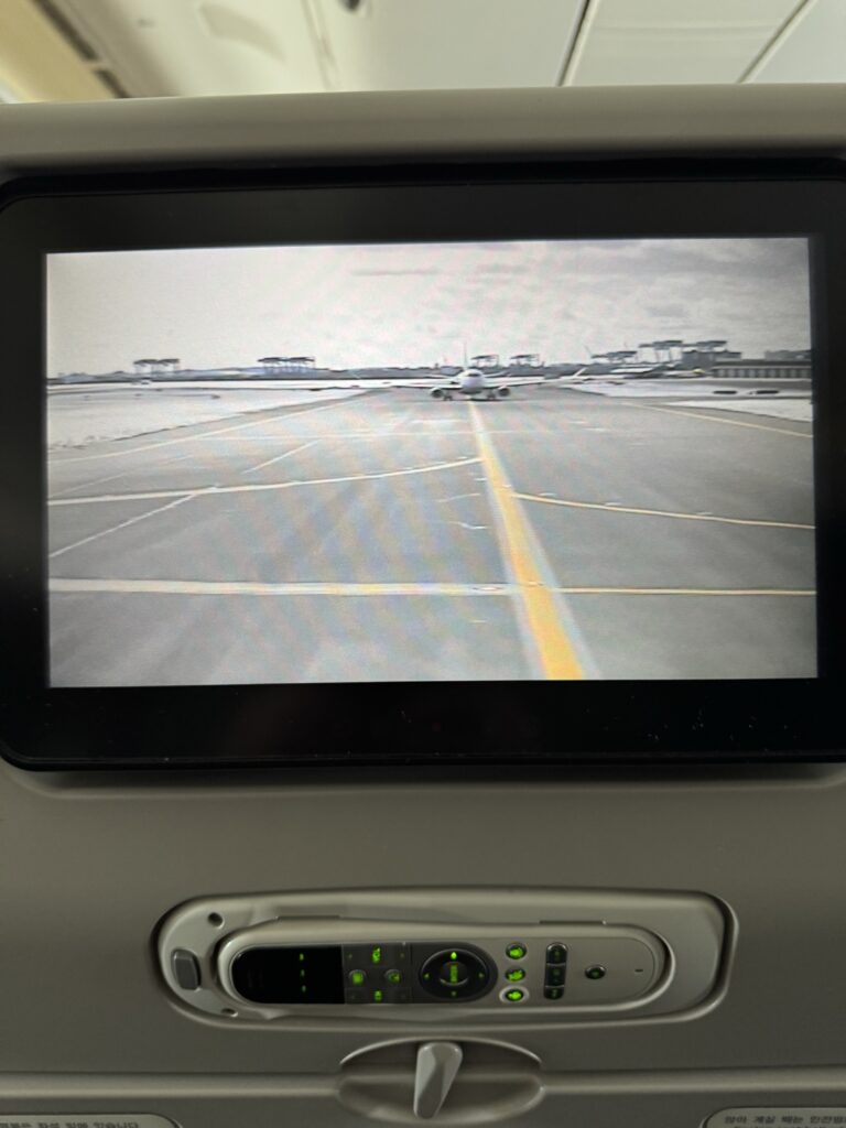 The seat back screen of an airplane seat showing the front facing camera on the aircraft. Another airplane can be seen queued in front.