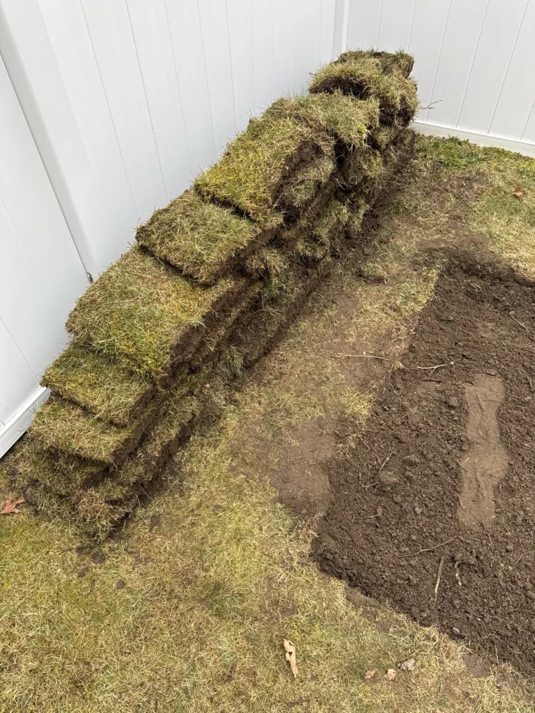 A pile of rectangular pieces of sod against a white vinyl fence with a square cut out in the grass.
