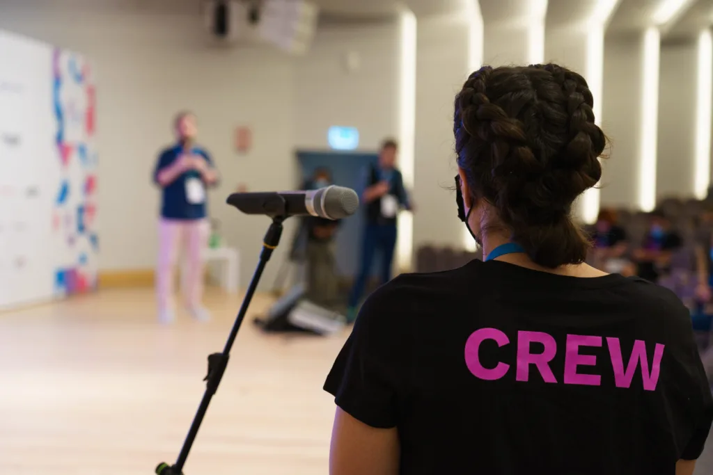 A person standing at a microphone in front of a stage asking a question. A person is visible but out of focus in the background.