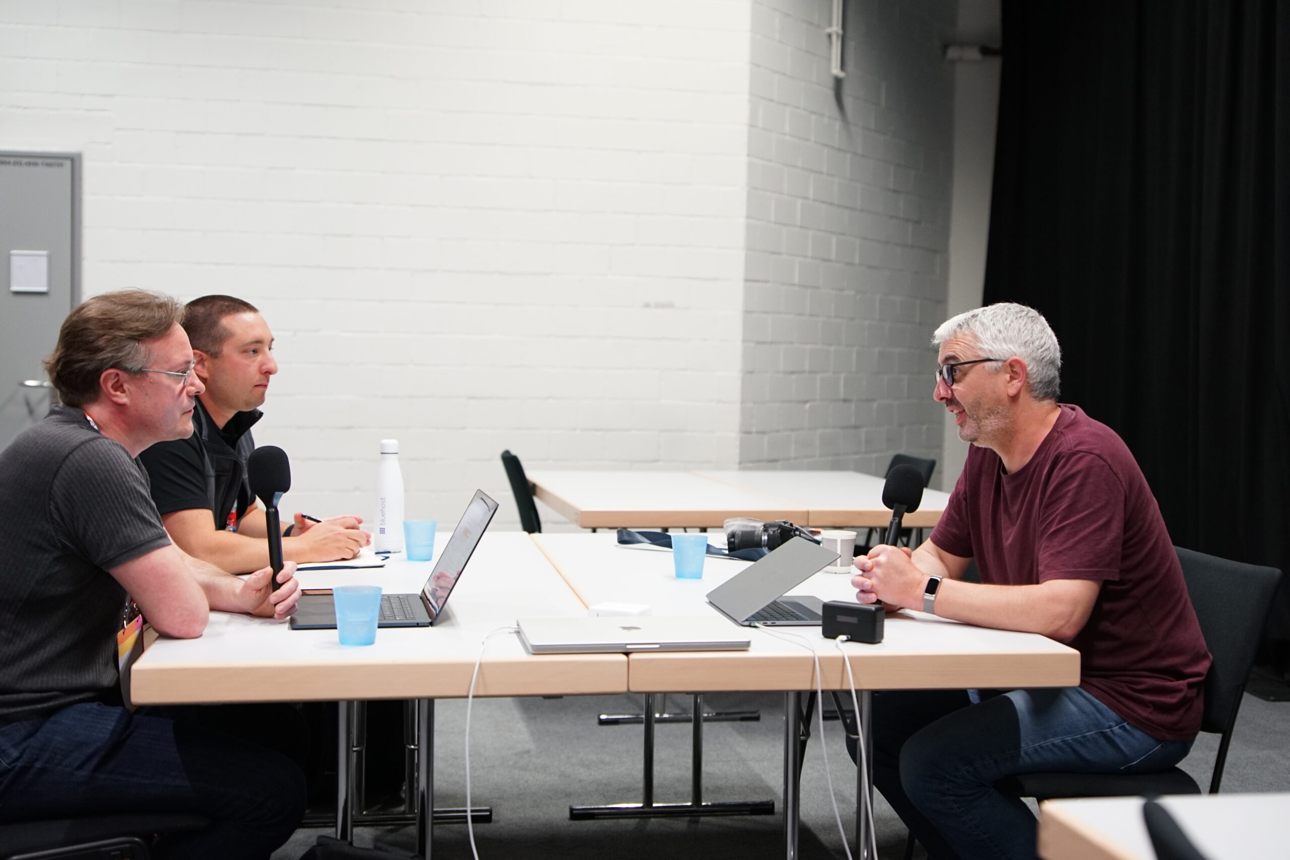 Jonathan Desrosiers and Joe Dolson sitting across from Nathan Wrigley recording an episode of the WP Tavern Jukebox podcast. There are laptops, cups of water, and notebooks on the table. They are holding microphones.