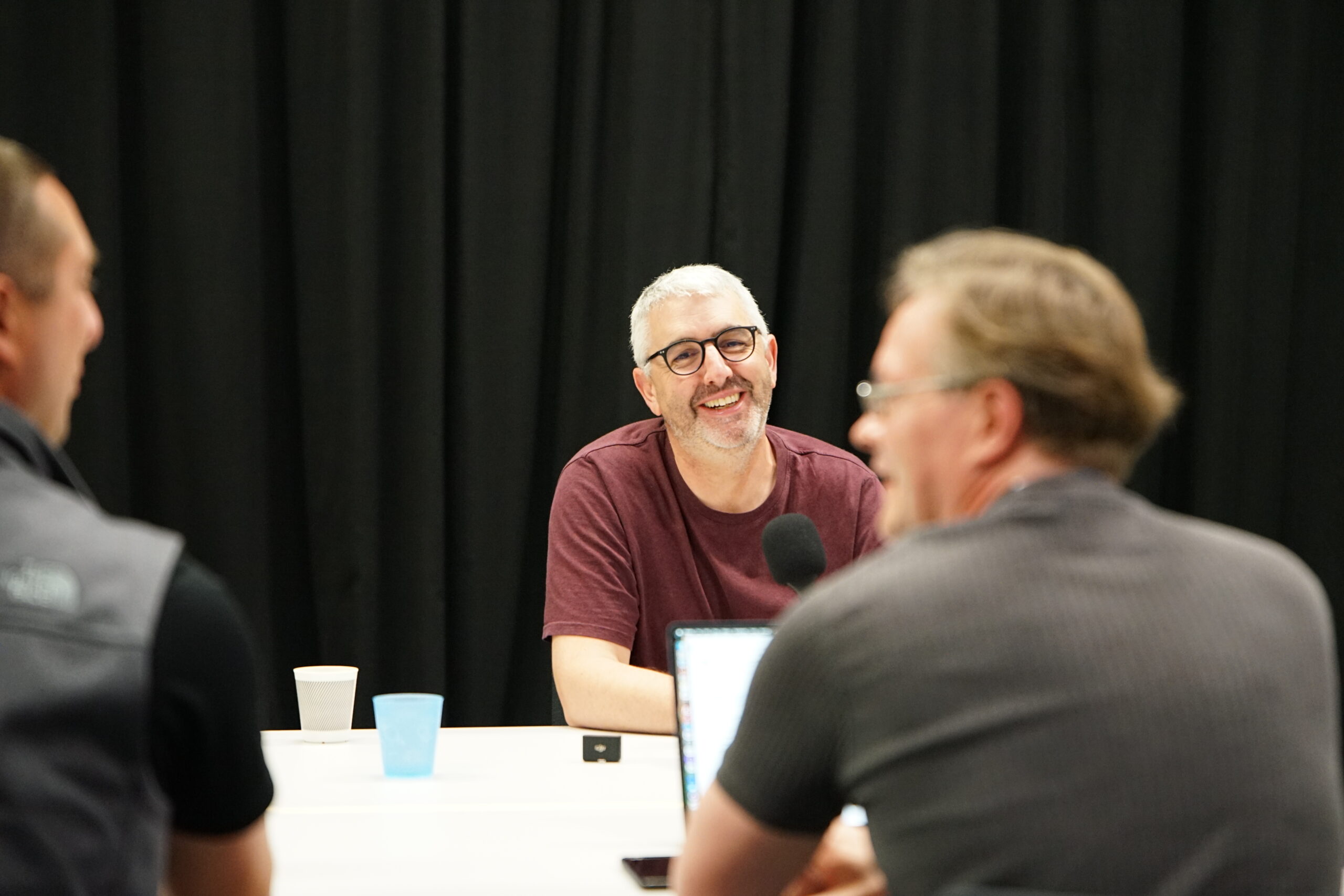 Nathan smiling at the camera form across the table. The camera is behind Jonathan's and Joe's backs.