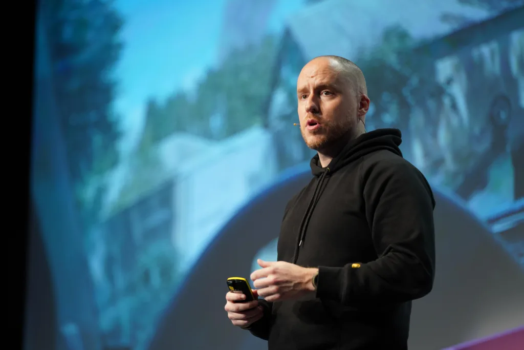Noel Tock on stage at WordCamp Europe. He's wearing a black hooded sweatshirt and is holding a remote control.