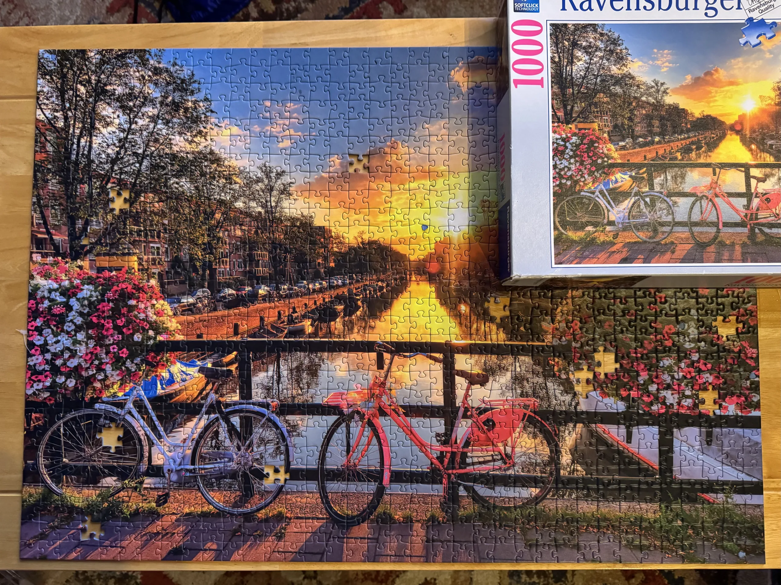 A completed Bicycles in Amsterdam 1,000 piece puzzle from Ravensbuger. The puzzle has bicycles leaning on the railing of a bridge over the Amstel river in Amsterdam with flowers on either side and a setting sun at the end of the river. The puzzle is on a light, natural wood coffee table.