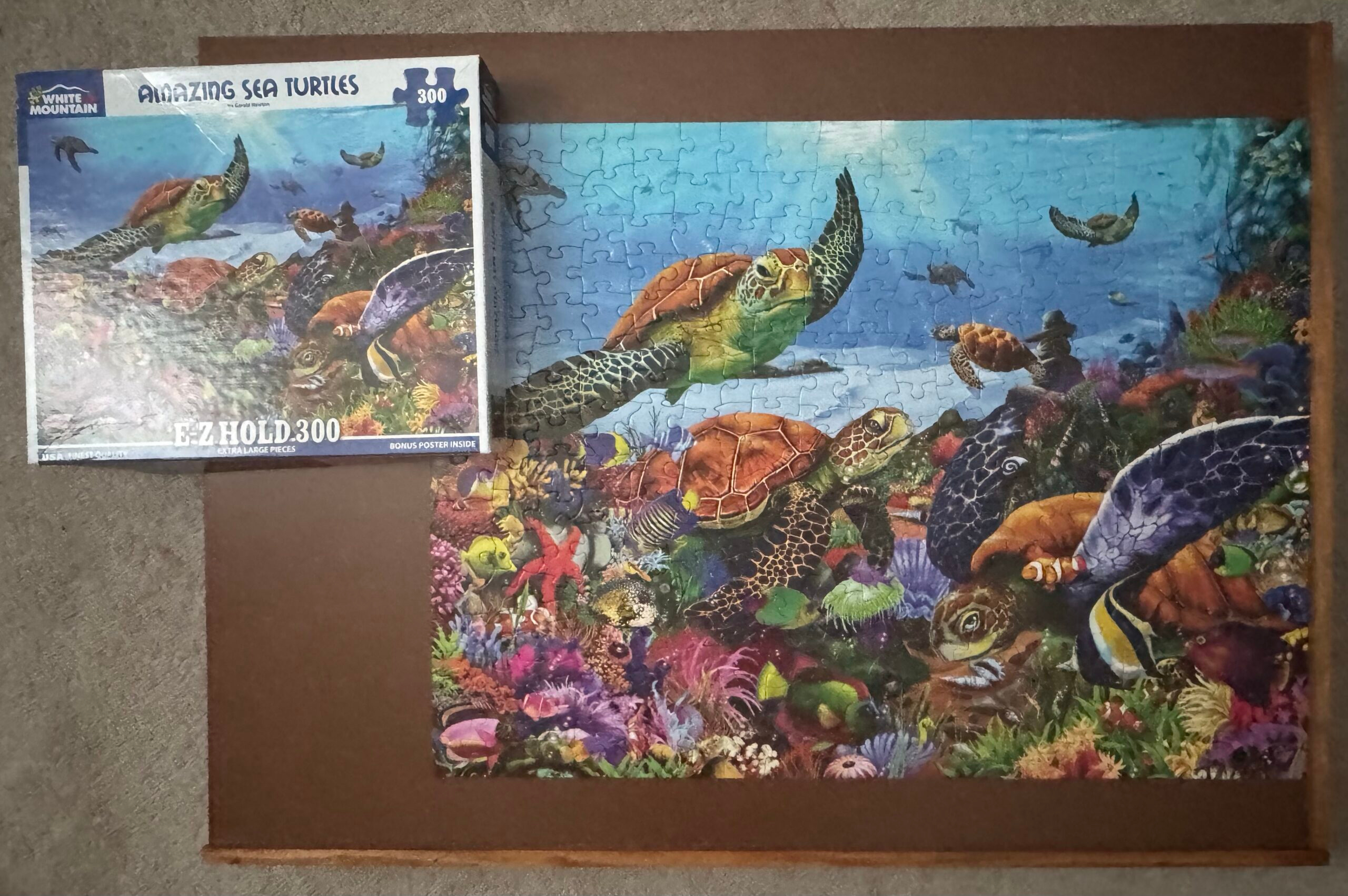 A completed Amazing Sea Turtles 300 piece puzzle from White Mountain Puzzles. The puzzle has several turtles swimming amongst other sea creatures near the bottom of the ocean. The puzzle is on a dark brown puzzle board placed on a tan carpet.
