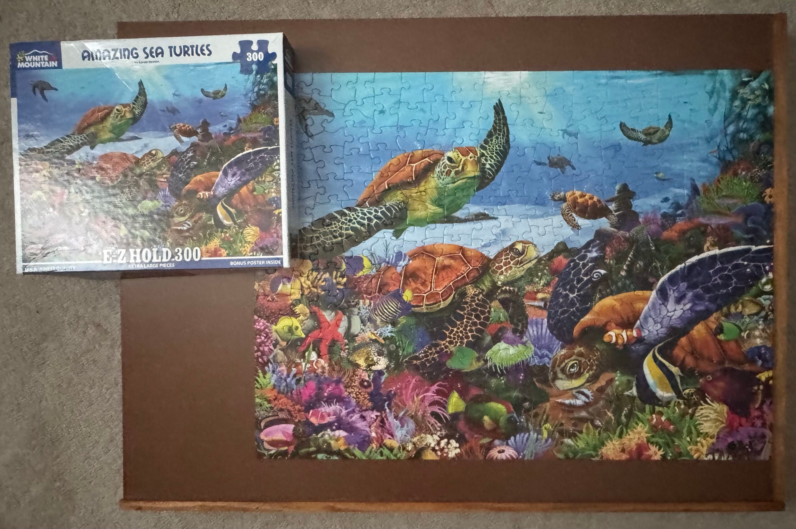A completed Amazing Sea Turtles 300 piece puzzle from White Mountain Puzzles. The puzzle has several turtles swimming amongst other sea creatures near the bottom of the ocean. The puzzle is on a dark brown puzzle board placed on a tan carpet.