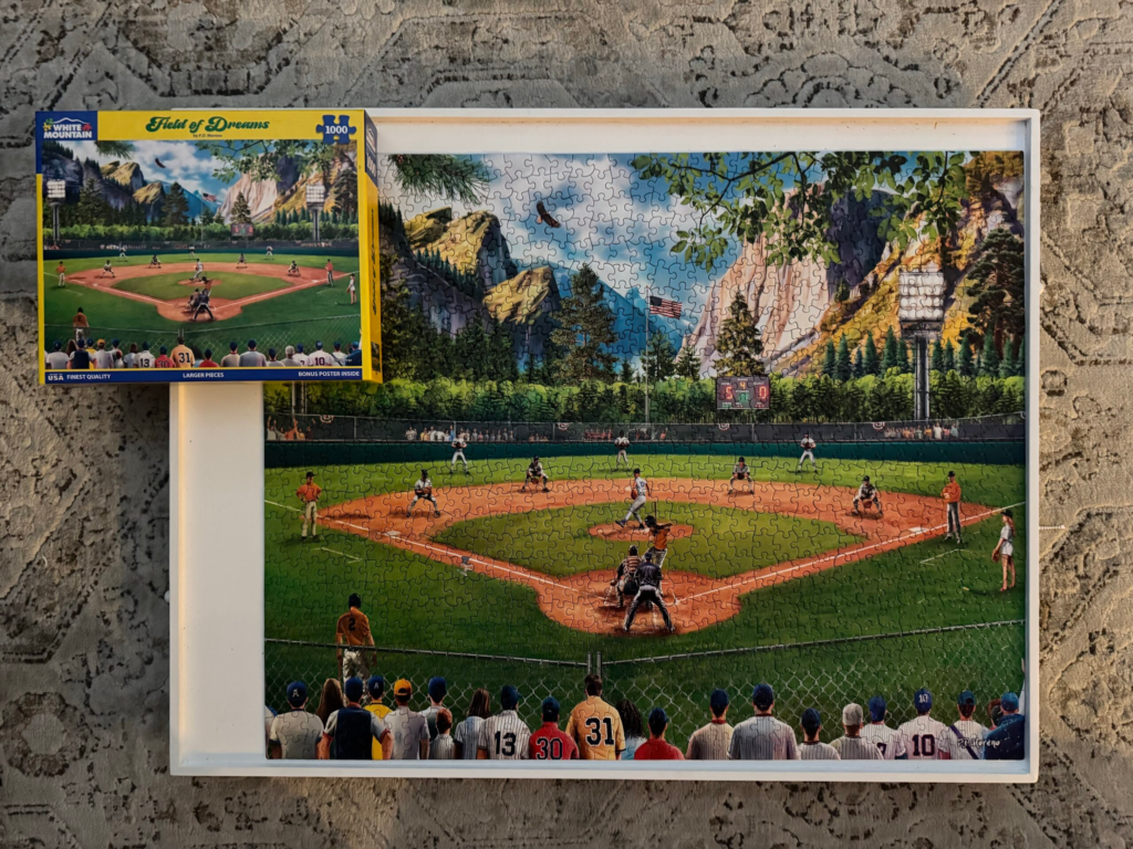 A completed AField of Dreams 1,000 piece jigsaw puzzle by White Mountain Puzzles. A baseball field surrounded by fans amongst a sprawling mountain backdrop is shown as the pitcher delivers a pitch to the batter. 