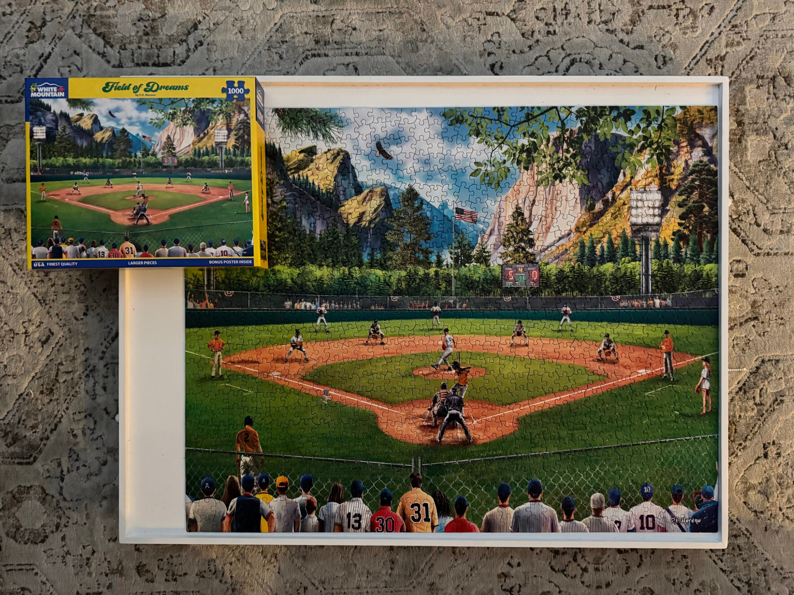 A completed AField of Dreams 1,000 piece jigsaw puzzle by White Mountain Puzzles. A baseball field surrounded by fans amongst a sprawling mountain backdrop is shown as the pitcher delivers a pitch to the batter.