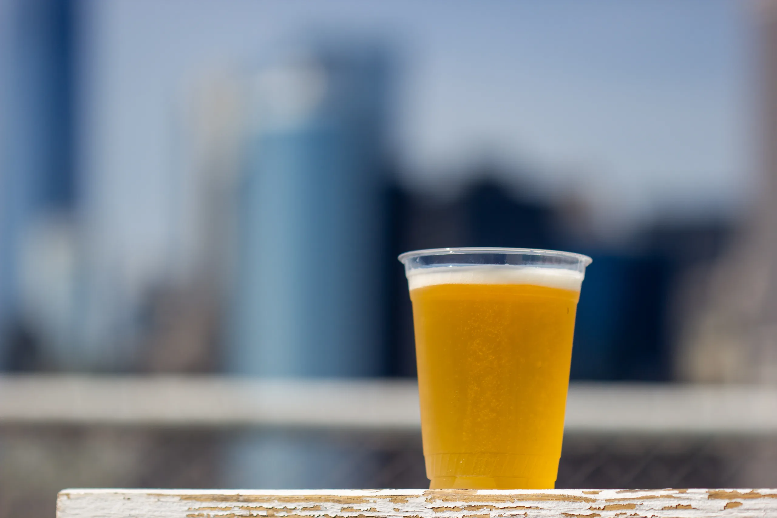Plastic glass of Beer, sitting on a board with white paint flaking off, a blurry city in the background.