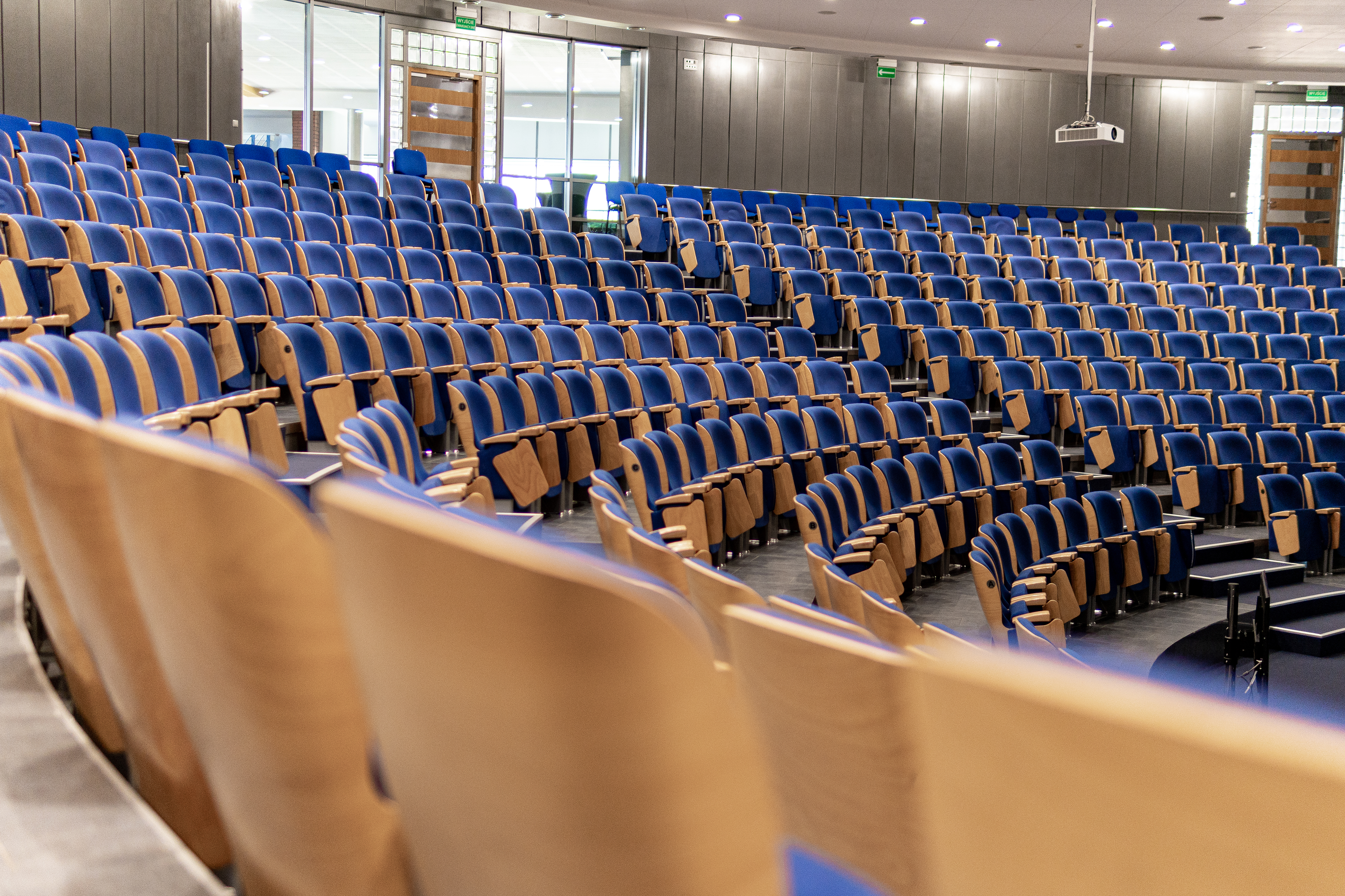 An empty auditorium in a university is furnished with wooden chairs that have blue cushions. The chairs are neatly arranged in rows, facing a stage or a podium that is not visible in the frame. The room is empty of people, and the lighting is relatively bright, daytime or artificial lighting. The overall atmosphere of the scene is academy.