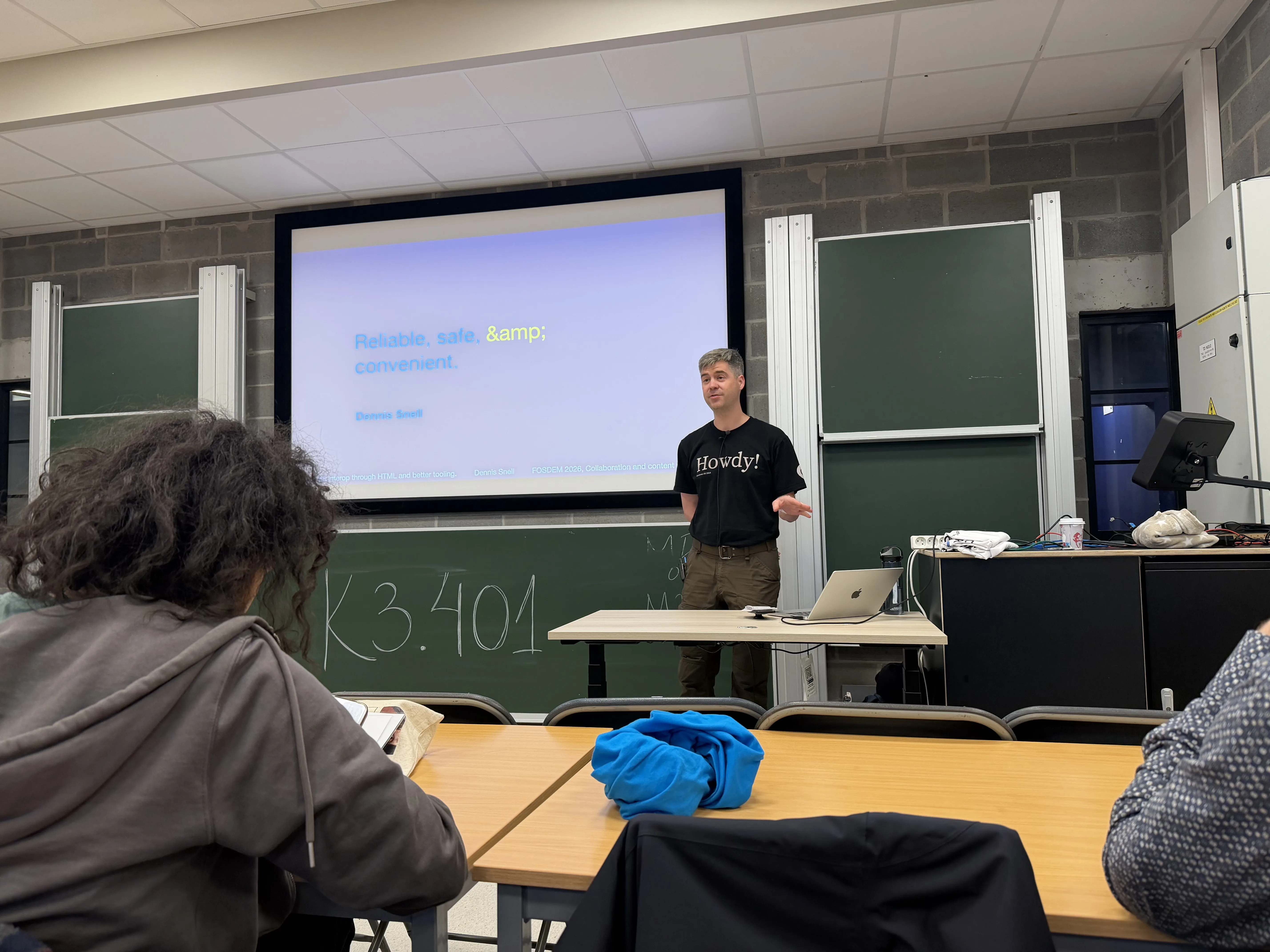 Dennis Snell standing in front of a projector screen presenting at FOSDEM 2026.